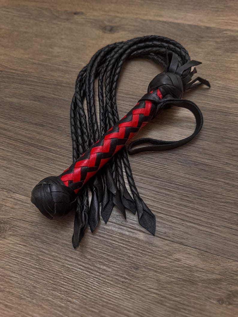 May include: A black leather whip with a red and black woven handle. The whip has a loop at the end of the handle and multiple black leather strands. The handle is approximately 6 inches long.