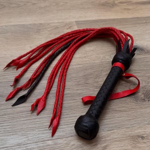 May include: A black and red leather whip with a braided handle and multiple red and black braided tails. The handle is textured and features a red wrist strap. The whip is lying on a wooden surface.
