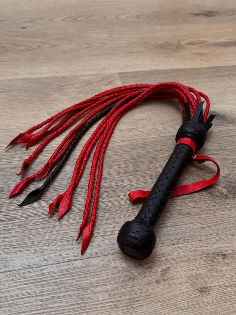 May include: A black and red leather whip with a braided handle and multiple red and black braided tails. The handle is textured and features a red wrist strap. The whip is lying on a wooden surface.