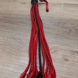 May include: A black and red leather whip with a black handle and a red loop. The whip is made of braided leather strands, with red strands and a few black strands. The whip is lying on a wooden surface.