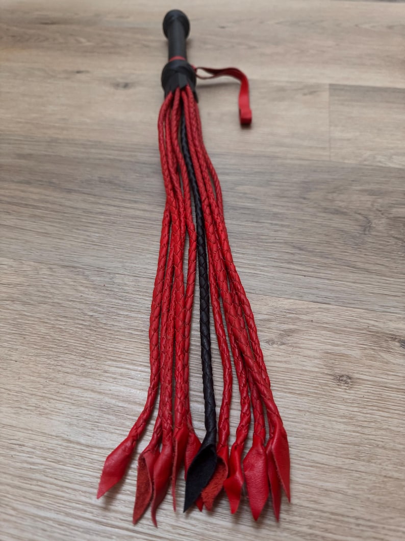 May include: A black and red leather whip with a black handle and a red loop. The whip is made of braided leather strands, with red strands and a few black strands. The whip is lying on a wooden surface.