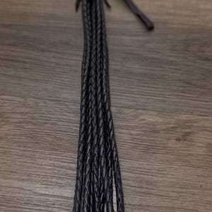 May include: A black leather whip with a braided handle and multiple braided strands. The handle is wrapped with red and black thread. The whip is displayed on a wooden surface.