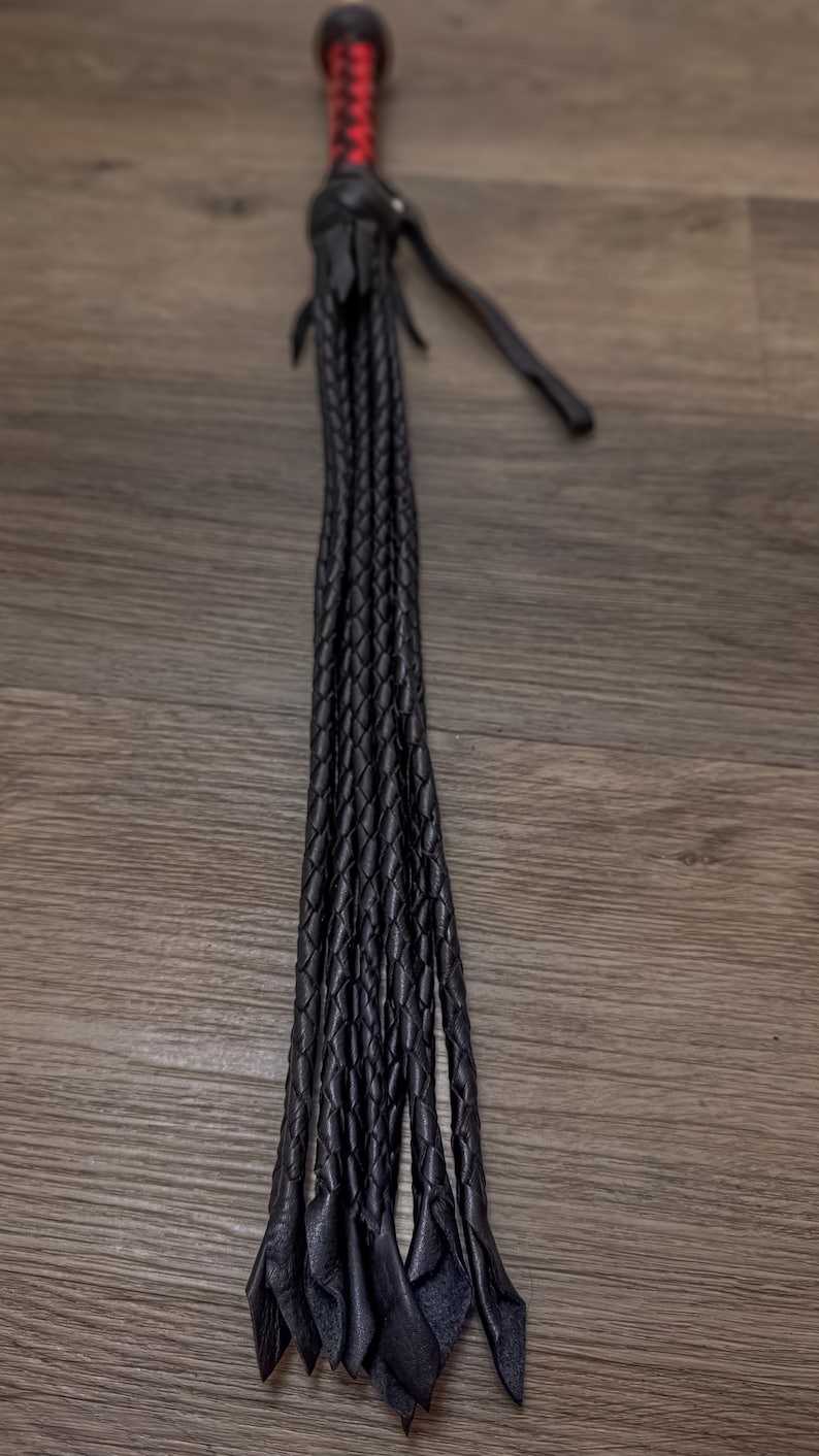 May include: A black leather whip with a braided handle and multiple braided strands. The handle is wrapped with red and black thread. The whip is displayed on a wooden surface.