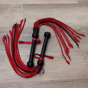 May include: Two black and red leather whips. Each whip has a textured black handle, a red strap, and long red and black braided strands. The whips are lying on a light wood surface.