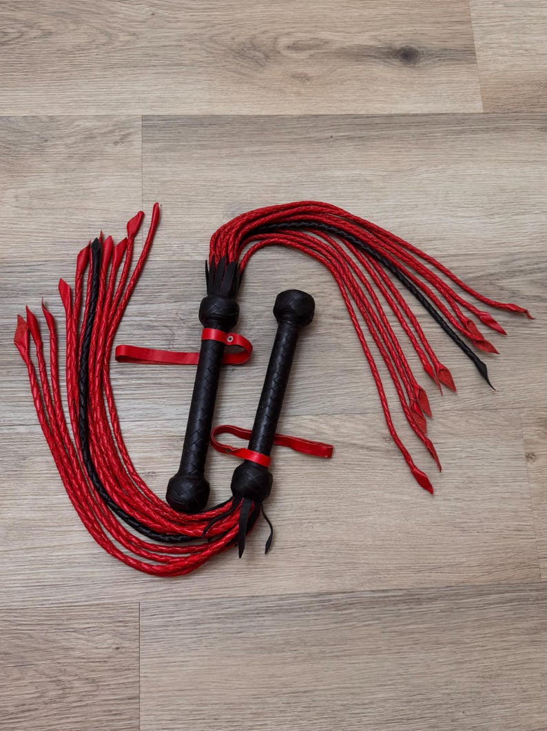May include: Two black and red leather whips. Each whip has a textured black handle, a red strap, and long red and black braided strands. The whips are lying on a light wood surface.