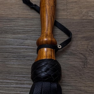 May include: A black leather flogger with a wooden handle. The handle is brown with a dark wood grain. The flogger has a braided leather strap and a tassel.