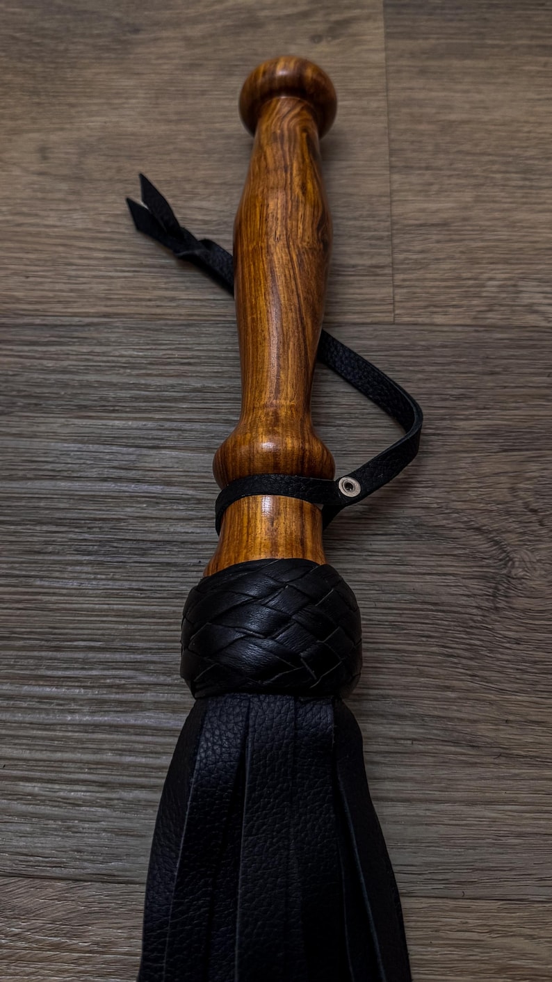 May include: A black leather flogger with a wooden handle. The handle is brown with a dark wood grain. The flogger has a braided leather strap and a tassel.