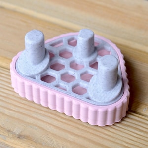 Puede incluir: Un objeto rosa y gris impreso en 3D con una base en forma de corazón. La base tiene un patrón de panal y tres postes cilíndricos. El borde de la base tiene un diseño festoneado. El objeto está sobre una superficie de madera.