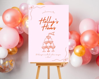 Puede incluir: Un cartel rosa y blanco con el texto "Bienvenido a Holly's Hens" en letra naranja. El cartel también presenta un dibujo de una torre de champán con el texto "28 días hasta que ella diga "¡Sí!" en naranja. El cartel está apoyado en un caballete de madera frente a un arco de globos.