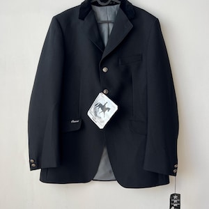 Könnte beinhalten: Eine schwarze Reitjacke mit Samtkragen, drei Knöpfen auf der Vorderseite und zwei Pattentaschen. Die Jacke ist mit einem Pikeur-Markenetikett mit Pferdegrafik sowie einem Pflegeetikett am Ärmel versehen und hängt an einem schwarzen Kleiderbügel.