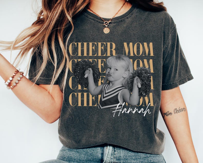 May include: Dark gray t-shirt with the words "CHEER MOM" in a gold color. The shirt features a black and white image of a young girl holding pom-poms, with the name "Hannah" in white script.