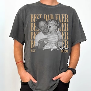Op de afbeelding: Donkergrijs t-shirt met een foto van een vader en kind. Het shirt heeft de tekst "BEST DAD EVER" in een herhalend patroon, met "Daddy & Sophia 2020" onder de foto. Het woord "EST." staat ook op het shirt.