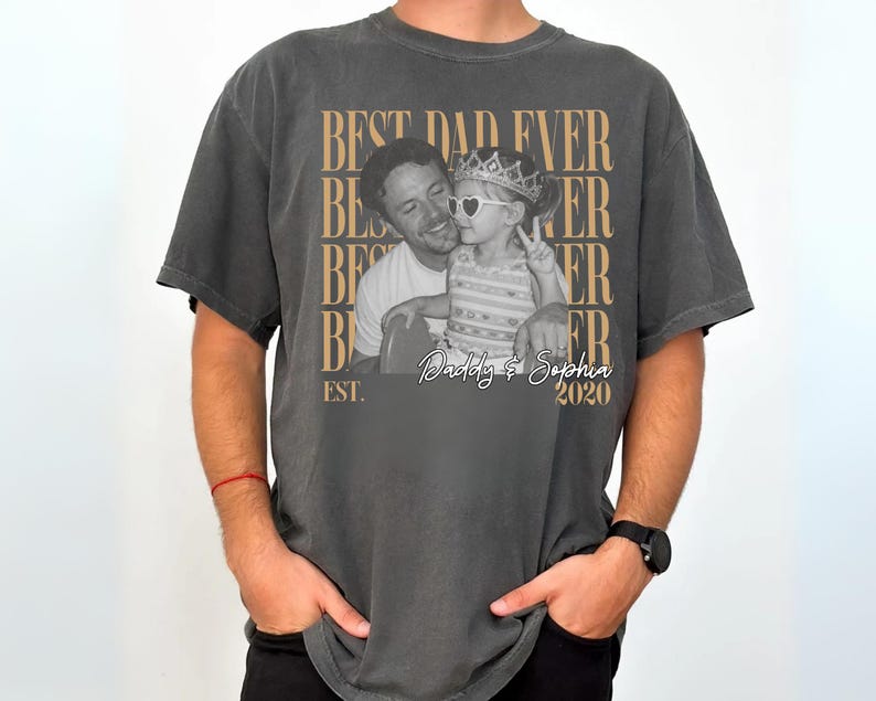 Op de afbeelding: Donkergrijs t-shirt met een foto van een vader en kind. Het shirt heeft de tekst "BEST DAD EVER" in een herhalend patroon, met "Daddy & Sophia 2020" onder de foto. Het woord "EST." staat ook op het shirt.