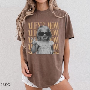 May include: An espresso-colored short-sleeve t-shirt featuring a graphic print of a child wearing sunglasses and sticking out their tongue while making a peace sign. The text reads ALEY'S MOM and EST. 2013 in a tan, serif font repeated in the background.