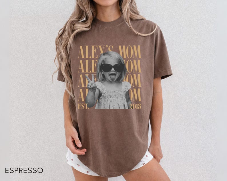 May include: An espresso-colored short-sleeve t-shirt featuring a graphic print of a child wearing sunglasses and sticking out their tongue while making a peace sign. The text reads ALEY'S MOM and EST. 2013 in a tan, serif font repeated in the background.