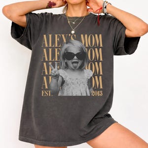 May include: A charcoal gray short-sleeve t-shirt featuring a black and white photograph of a child wearing sunglasses and making a peace sign. The text ALEY'S MOM is repeated in tan lettering behind the child, with EST. 2013 printed at the bottom.