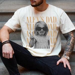 K&ouml;nnte beinhalten: Cremefarbenes T-Shirt mit einem Schwarz-Wei&szlig;-Foto eines Kindes mit Sonnenbrille und Peace-Zeichen. Der Text "ALEX'S DAD" ist vertikal gedruckt, mit "EST 2018" am unteren Rand.