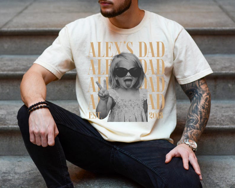 K&ouml;nnte beinhalten: Cremefarbenes T-Shirt mit einem Schwarz-Wei&szlig;-Foto eines Kindes mit Sonnenbrille und Peace-Zeichen. Der Text "ALEX'S DAD" ist vertikal gedruckt, mit "EST 2018" am unteren Rand.