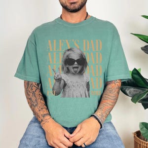 K&ouml;nnte beinhalten: Ein t&uuml;rkisgr&uuml;nes T-Shirt mit einem Schwarz-Wei&szlig;-Foto eines Kindes mit Sonnenbrille und herausgestreckter Zunge. Der Text "ALEX'S DAD" ist oberhalb und unterhalb des Fotos aufgedruckt, mit "EST 2018" darunter.