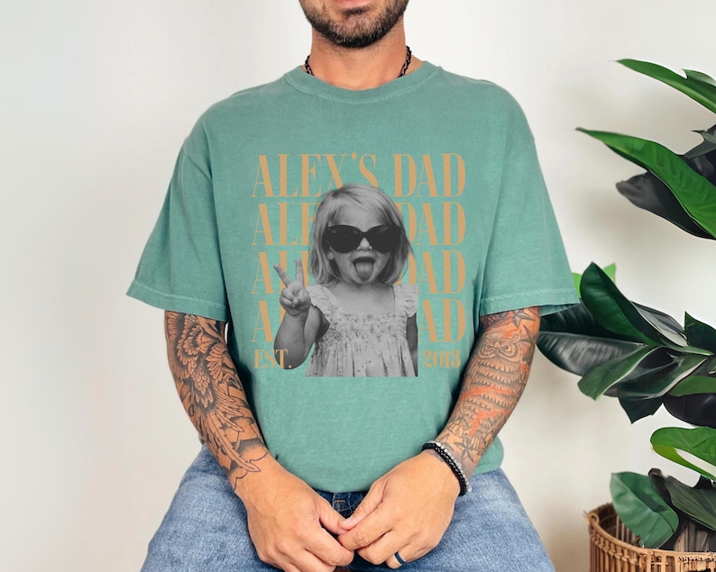 K&ouml;nnte beinhalten: Ein t&uuml;rkisgr&uuml;nes T-Shirt mit einem Schwarz-Wei&szlig;-Foto eines Kindes mit Sonnenbrille und herausgestreckter Zunge. Der Text "ALEX'S DAD" ist oberhalb und unterhalb des Fotos aufgedruckt, mit "EST 2018" darunter.