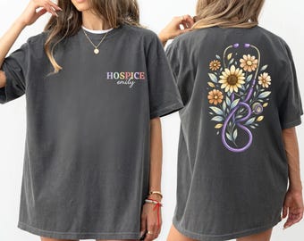 May include: A charcoal gray t-shirt featuring the text "HOSPICE emily" in multicolored letters on the front left chest. The back displays a large graphic of a purple stethoscope intertwined with yellow and orange sunflowers and green leaves.