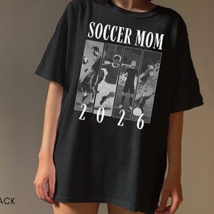 May include: Black t-shirt with the text "SOCCER MOM 2026" in white. The design includes a series of black and white images of soccer players. The shirt is a casual, short-sleeved style.