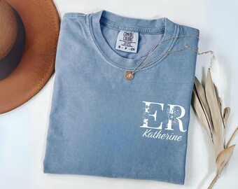 May include: A blue short-sleeve t-shirt with a crew neckline featuring a white graphic on the chest that displays the letters ER with floral details and the name Katherine written below in a script font. The shirt has a Comfort Colors brand label.