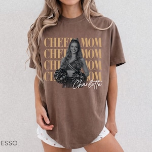 May include: Espresso-colored t-shirt with the words "CHEER MOM" repeated vertically in a gold color. A black and white photo of a cheerleader is in the center, with the name "Charlotte" in cursive below. The shirt is a casual style.