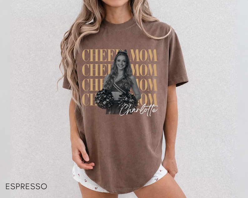 May include: Espresso-colored t-shirt with the words "CHEER MOM" repeated vertically in a gold color. A black and white photo of a cheerleader is in the center, with the name "Charlotte" in cursive below. The shirt is a casual style.