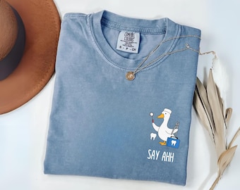 May include: A blue short-sleeved t-shirt with a graphic of a white goose dressed as a dentist holding a dental mirror and pliers next to a blue medical bag and a tooth. The text "SAY AHH" is printed below the graphic in white lettering.