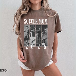 May include: Espresso-colored t-shirt with the text "SOCCER MOM 2026" in white. The shirt features a black and white collage of soccer players. The shirt is a short-sleeved, crew neck style.