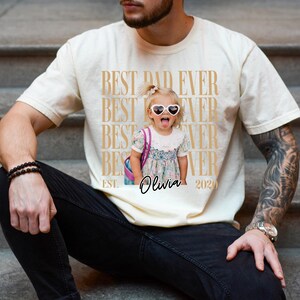 May include: Cream-colored t-shirt with the text "BEST DAD EVER" repeated vertically in a gold color. A photo of a young girl wearing heart-shaped sunglasses is printed on the shirt, with the name "Olivia" and the year "2020" below.