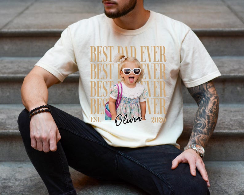 May include: Cream-colored t-shirt with the text "BEST DAD EVER" repeated vertically in a gold color. A photo of a young girl wearing heart-shaped sunglasses is printed on the shirt, with the name "Olivia" and the year "2020" below.