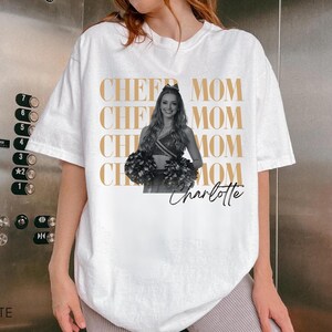 May include: White t-shirt with the words "CHEER MOM" repeated in a gold color. A black and white image of a cheerleader holding pom-poms is on the shirt, with the name "Charlotte" in script below.
