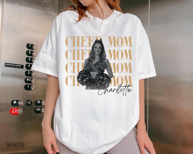 May include: White t-shirt with the words "CHEER MOM" repeated in a gold color. A black and white image of a cheerleader holding pom-poms is on the shirt, with the name "Charlotte" in script below.