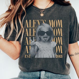 May include: A charcoal gray t-shirt featuring a black and white photograph of a child wearing sunglasses and making a peace sign. The text reads ALEY'S MOM repeated in gold, with EST. 2013 at the bottom. The shirt has a crew neck and short sleeves.