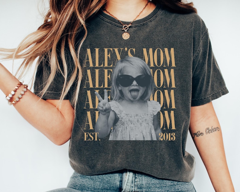 May include: A charcoal gray t-shirt featuring a black and white photograph of a child wearing sunglasses and making a peace sign. The text reads ALEY'S MOM repeated in gold, with EST. 2013 at the bottom. The shirt has a crew neck and short sleeves.