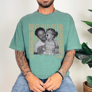 Op de afbeelding: Een zeegroen t-shirt met een zwart-wit foto van een man en een kind. De tekst "BEST DAD EVER" is verticaal in een herhalend patroon gedrukt. De foto is gelabeld "Daddy & Sophia".