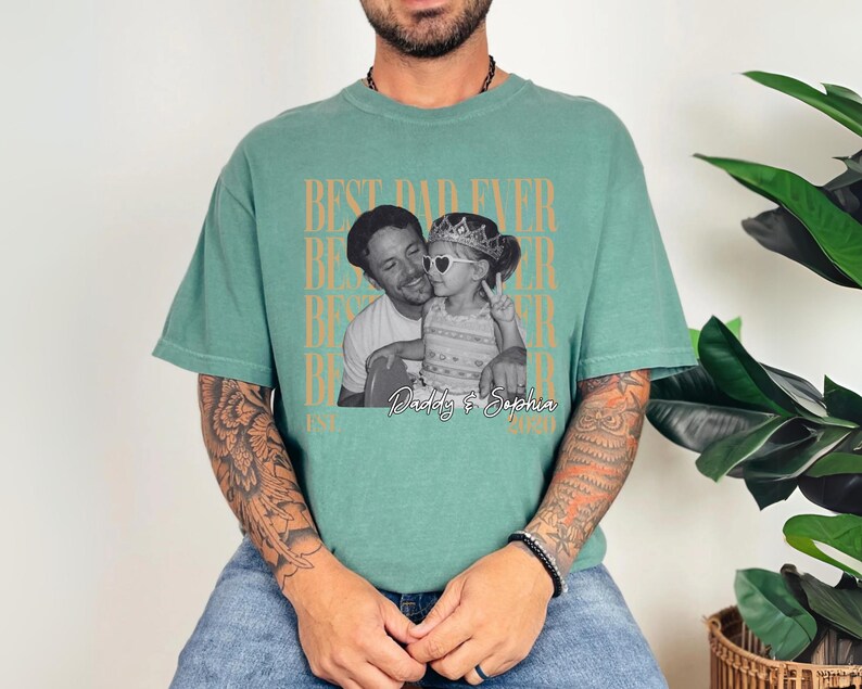 Op de afbeelding: Een zeegroen t-shirt met een zwart-wit foto van een man en een kind. De tekst "BEST DAD EVER" is verticaal in een herhalend patroon gedrukt. De foto is gelabeld "Daddy & Sophia".