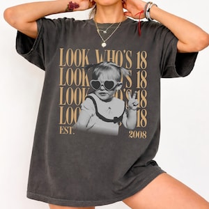 May include: A charcoal gray short-sleeve t-shirt featuring a graphic print of a child wearing heart-shaped sunglasses and making a peace sign. The text "LOOK WHO'S 18" is repeated in a tan font, with "EST. 2008" printed at the bottom of the design.