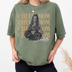 May include: Olive green t-shirt with the words "CHEE MOM" repeated in a gold color. A black and white photo of a cheerleader is in the center, with the name "Charlotte" in script below.