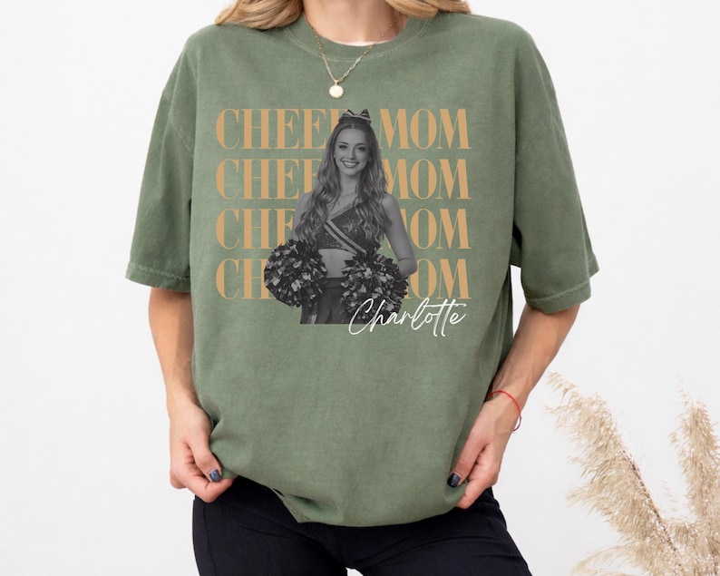 May include: Olive green t-shirt with the words "CHEE MOM" repeated in a gold color. A black and white photo of a cheerleader is in the center, with the name "Charlotte" in script below.