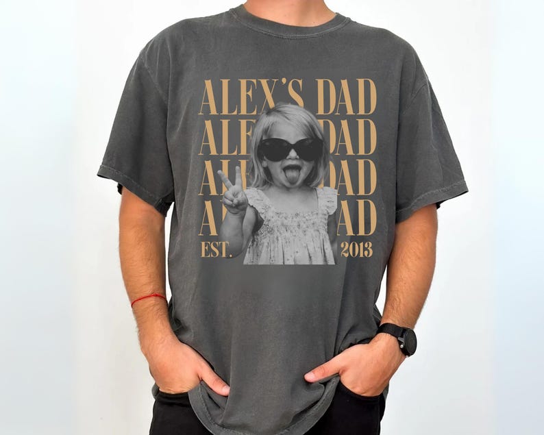 K&ouml;nnte beinhalten: Dunkelgraues T-Shirt mit einem Schwarz-Wei&szlig;-Foto eines Kindes mit Sonnenbrille und Peace-Zeichen. Der Text "ALEX'S DAD" ist &uuml;ber dem Foto aufgedruckt, darunter "EST. 2013".