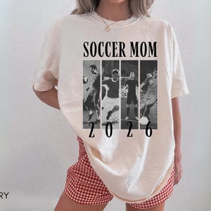 May include: Ivory-colored t-shirt with the text "SOCCER MOM 2026" in black. The design includes a collage of black and white soccer action shots. The shirt is a casual, oversized fit.