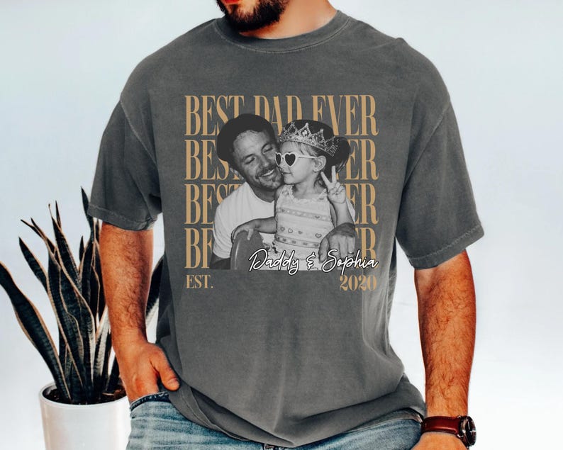 Op de afbeelding: Grijze t-shirt met een zwart-wit foto van een man en een kind. De tekst "BEST DAD EVER" staat boven de foto, met "Daddy & Sophia 2020" eronder. Het shirt heeft een casual stijl.