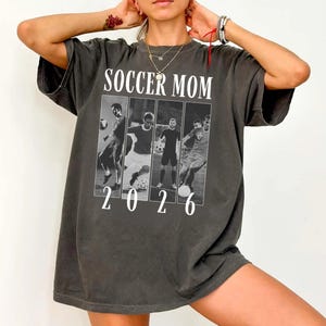 May include: Dark gray t-shirt with the words "SOCCER MOM" above a black and white image of soccer players. The number "2026" is printed below the image. The shirt has short sleeves and a relaxed fit.