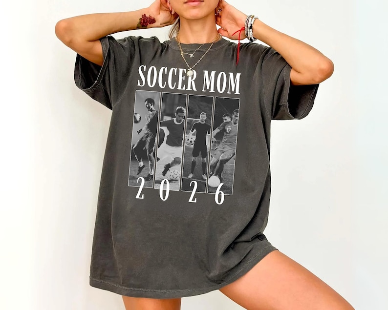 May include: Dark gray t-shirt with the words "SOCCER MOM" above a black and white image of soccer players. The number "2026" is printed below the image. The shirt has short sleeves and a relaxed fit.
