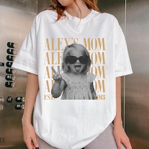 May include: A white short-sleeve t-shirt featuring a black and white photograph of a child wearing sunglasses, making a peace sign and sticking out their tongue. The text "ALEX'S MOM" is repeated in gold, with "EST. 2013" printed at the bottom.