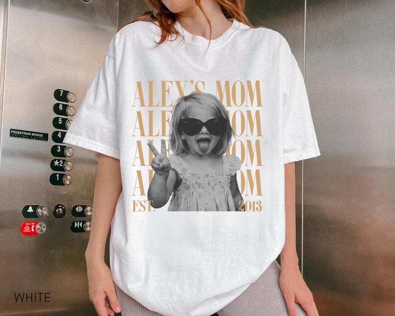 May include: A white short-sleeve t-shirt featuring a black and white photograph of a child wearing sunglasses, making a peace sign and sticking out their tongue. The text "ALEX'S MOM" is repeated in gold, with "EST. 2013" printed at the bottom.