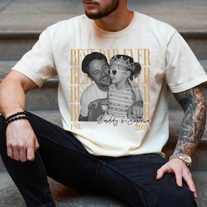 Op de afbeelding: Cr&egrave;mekleurig T-shirt met een zwart-wit foto van een man en een kind. Het shirt heeft de tekst "BEST DAD EVER" verticaal herhaald en "Daddy & Sophia 2020" onder de foto. Het shirt wordt door een persoon gedragen.